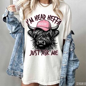 May include: White t-shirt with a black and white illustration of a Highland cow wearing a pink hat that says "Bossy". The text "I'm Head Heffa Just Ask Me" is printed below the cow.