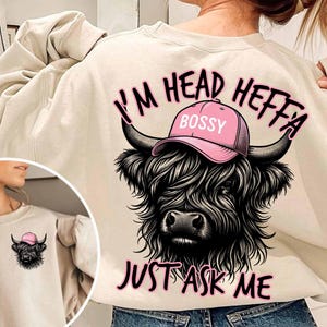 May include: A cream colored sweatshirt with a black and white illustration of a Highland cow wearing a pink hat that says "Bossy". The text "I'm Head Heffa Just Ask Me" is printed in pink on the sweatshirt.