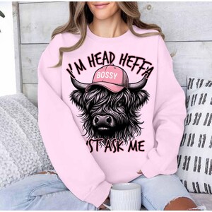 May include: Pink sweatshirt with a black and white Highland cow wearing a pink hat that says "Bossy" and the text "I'm Head Heffa Don't Ask Me".