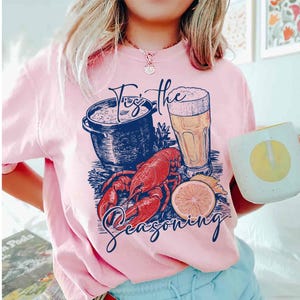 May include: A pink t-shirt with a graphic design of a pot of seafood, a glass of beer, a lobster, and lemons. The text on the shirt reads "Tis the Seasoning".