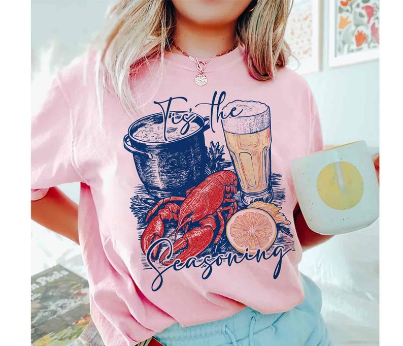 May include: A pink t-shirt with a graphic design of a pot of seafood, a glass of beer, a lobster, and lemons. The text on the shirt reads "Tis the Seasoning".