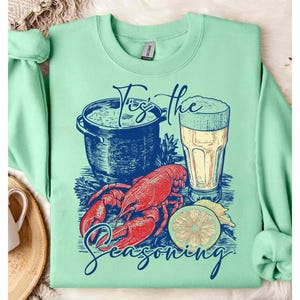 May include: A green sweatshirt with a graphic design of a lobster, a pot of seafood, a lemon, and a glass of beer. The text "Tis the Seasoning" is printed on the sweatshirt.