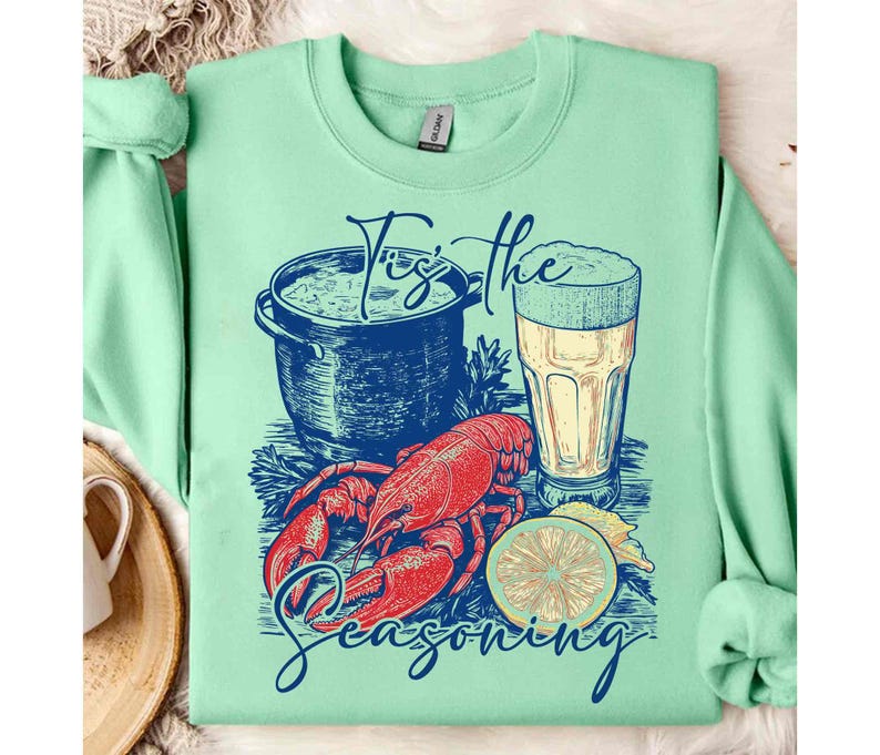 May include: A green sweatshirt with a graphic design of a lobster, a pot of seafood, a lemon, and a glass of beer. The text "Tis the Seasoning" is printed on the sweatshirt.