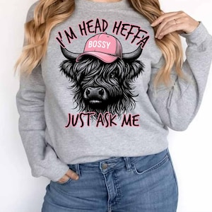 May include: Gray sweatshirt with a black and white illustration of a Highland cow wearing a pink hat that says "Bossy". The text "I'm Head Heffa Just Ask Me" is printed on the sweatshirt.