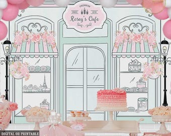 May include: A decorative backdrop featuring a cafe scene with pink and white balloons. The cafe has a sign that reads "Rosey's Cafe" and is decorated with pink and white flowers. A pink and white cake is on display.