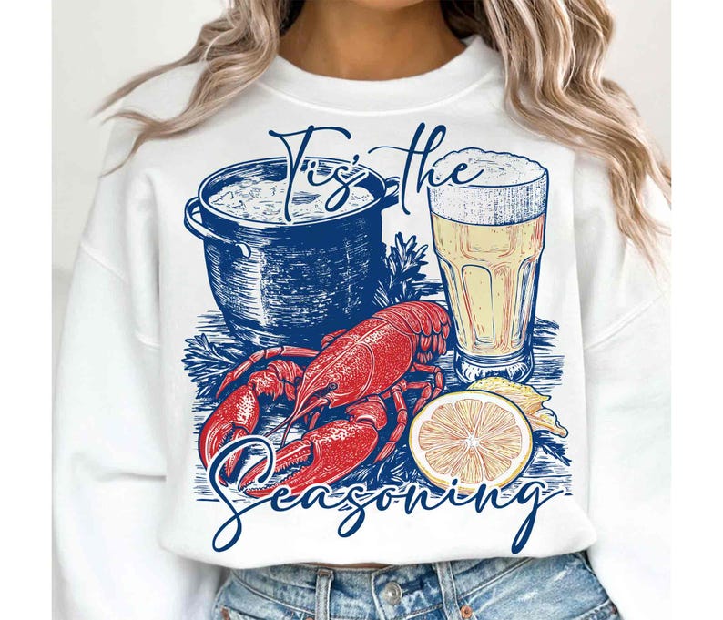 May include: White sweatshirt with a red lobster, a pot of seafood, a glass of beer, and lemons. The text "Tis the Seasoning" is printed on the sweatshirt.