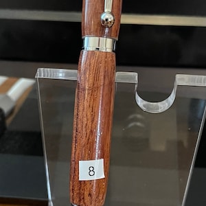 May include: A brown wooden pen with silver accents and a stylus tip. The pen is numbered 8 and is displayed on a clear acrylic stand.