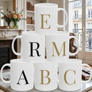 May include: A stack of six white ceramic mugs arranged in a pyramid. Each mug features a large, centered serif letter in either gold or black. The top mug displays an E, the middle row shows an R and an M, and the bottom row shows an A, B, and C.