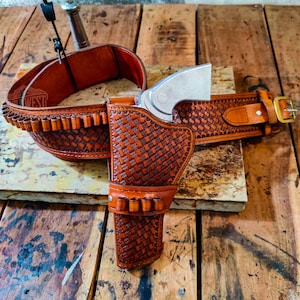 Può includere: Un set di fondina e cintura per pistola in pelle marrone con un motivo a trama di vimini. Il set include una fondina, una cintura e un porta cartucce. La cintura ha una fibbia color oro. La parola "WEST" è visibile sulla cintura.