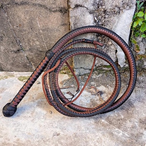 May include: A braided whip with a handle and a long, coiled lash. The whip features a pattern of alternating brown and black sections. The handle has a diamond pattern, and the lash is coiled in a circular shape. The whip is lying on a concrete surface.