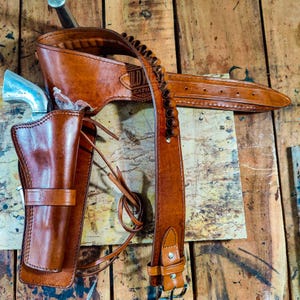 May include: A brown leather gun holster and belt set. The set includes a holster, a belt with bullet loops, and a shoulder strap. The leather is a rich, warm brown color. The set is designed for a revolver.