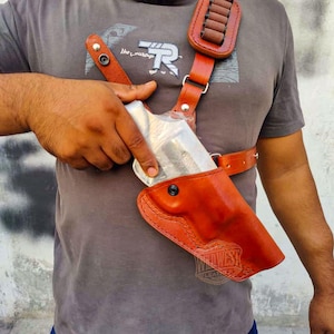 May include: A brown leather shoulder holster with a matching ammo holder. The holster is designed to be worn across the chest and features a stitched design. A silver handgun is partially visible in the holster.