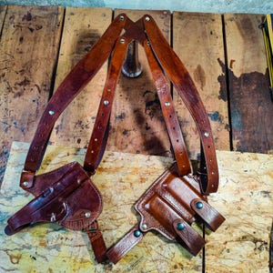 Puede incluir: Una funda de hombro de cuero marrón con dos bolsillos para pistolas y correas ajustables. La funda está diseñada para llevarse sobre los hombros y la espalda, con los bolsillos para pistolas colocados debajo de los brazos. El cuero es de un color marrón oscuro intenso.