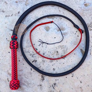 May include: A black and red whip with a braided handle and a long, flexible lash. The handle is red with black accents, and the lash is black. The whip is coiled on a concrete surface.