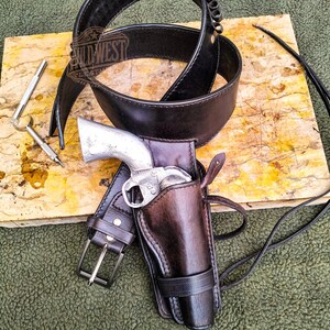 Op de afbeelding: Een zwarte leren riem en een bruin leren holster met een zilverkleurig revolver. Het holster is bevestigd aan een bijpassende riem. De afbeelding bevat ook een zwarte leren nekriem en twee zilveren gereedschappen.