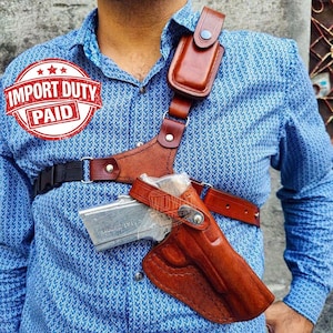 Peut inclure: Un système d'étui d'épaule en cuir marron comprenant une pochette moulée pour arme à feu, une pochette accessoire secondaire sur la bandoulière et des sangles réglables en cuir et en nylon noir, porté sur une chemise bleue à motifs.