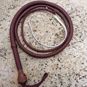 May include: A braided burgundy whip with a white handle and a loop at the end. The whip is coiled on a textured, light-colored surface. The handle is wrapped with a decorative pattern.