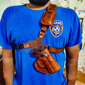 May include: A brown leather shoulder holster with a matching magazine pouch. The holster has a vertical design with adjustable straps and a secure fit. The leather is a rich, medium brown colour, and the stitching is visible. The holster is designed to be worn under the arm.