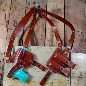 Puede incluir: Una funda de hombro de cuero marrón con una correa de cinturón a juego. La funda está diseñada para sujetar una pistola, con un objeto azul claro en el interior. El cuero está cosido y remachado para mayor durabilidad.