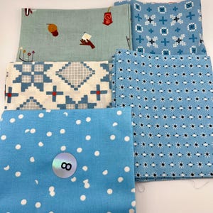 May include: Five folded fabric squares featuring various patterns. One is light green with small icons, one is blue with white cross-stitch motifs, one is cream with blue and gray geometric shapes, one is light blue with black starbursts, and one is bright blue with white polka dots and a circular sticker with the number 8.