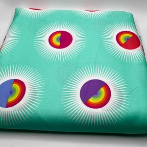 May include: A folded piece of fabric with a repeating pattern of large, stylized sunbursts. Each sunburst features a rainbow-colored arc within a magenta or purple circle, surrounded by white rays on a teal background.