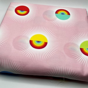 May include: Pink fabric with a repeating pattern of colorful sunburst designs. Each sunburst features a rainbow-colored arc within a yellow, red, green, or purple circle, surrounded by white radiating lines. The fabric is folded.