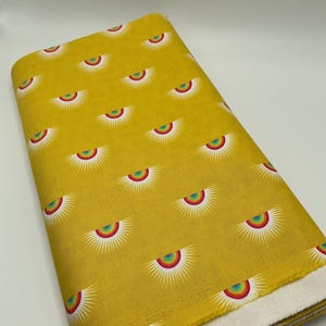 May include: A folded fabric bolt featuring a repeating pattern of stylized suns with rainbow arcs on a mustard yellow background. The suns have white rays emanating from the rainbow arcs. The fabric has a white selvedge edge.