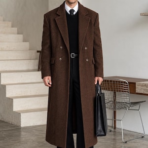 May include: A long, double-breasted brown wool coat with notched lapels and four buttons, worn over a black sweater, a white collared shirt with a black tie, and black trousers. A black leather tote bag is held at the side with black shoes.