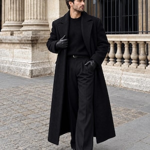 May include: A long black wool overcoat worn over a black ribbed sweater and black pleated trousers. The outfit includes black leather gloves and black leather boots. The coat features wide lapels and a structured fit, creating a monochromatic look.