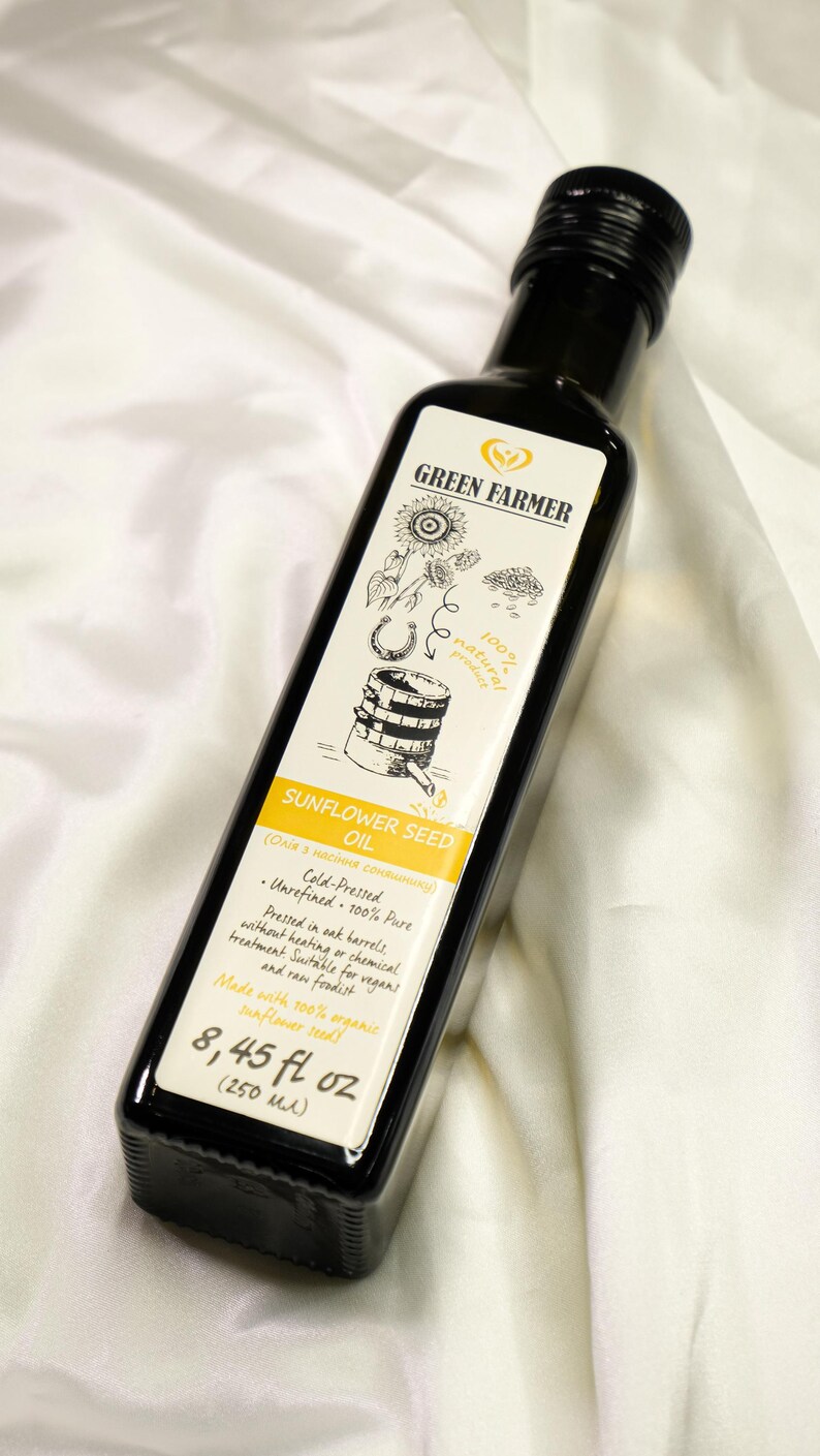 K&ouml;nnte beinhalten: Eine dunkle Glasflasche mit Green Farmer Sonnenblumenkern&ouml;l. Das Etikett zeigt Illustrationen von Sonnenblumen, einem Hufeisen und einem Fass. Die Flasche enth&auml;lt 250 ml kaltgepresstes, unraffiniertes &Ouml;l.