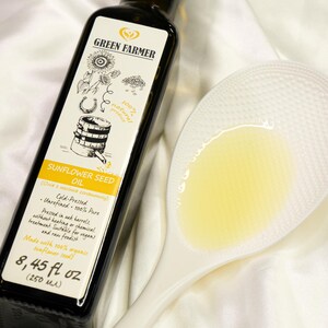 K&ouml;nnte beinhalten: Eine dunkle Glasflasche Sonnenblumen&ouml;l von Green Farmer mit wei&szlig;em Etikett, neben einem wei&szlig;en L&ouml;ffel mit &Ouml;l gef&uuml;llt. Das Etikett enth&auml;lt Text und Illustrationen. Die Flasche enth&auml;lt 250 ml &Ouml;l.