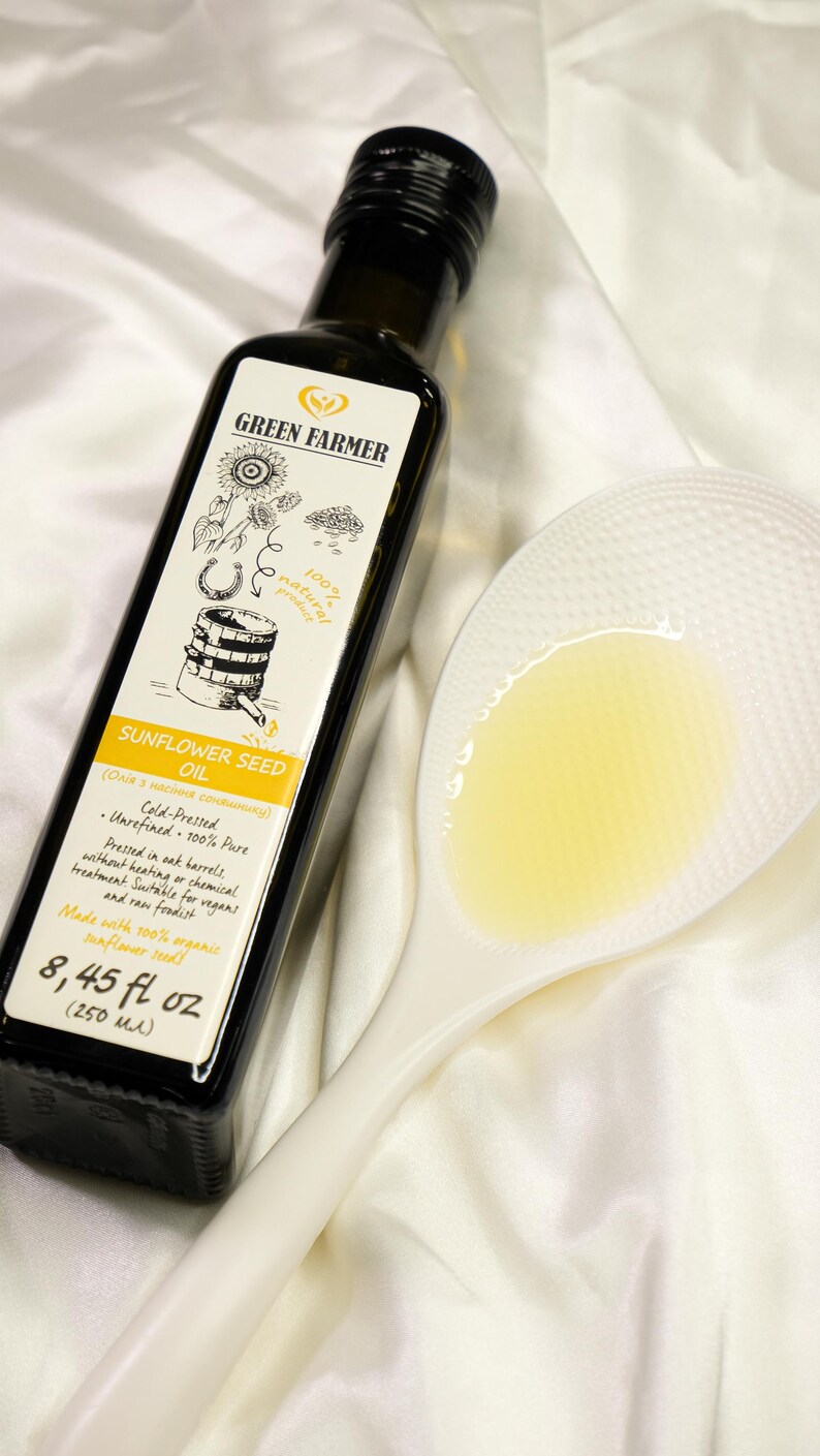 K&ouml;nnte beinhalten: Eine dunkle Glasflasche Sonnenblumen&ouml;l von Green Farmer mit wei&szlig;em Etikett, neben einem wei&szlig;en L&ouml;ffel mit &Ouml;l gef&uuml;llt. Das Etikett enth&auml;lt Text und Illustrationen. Die Flasche enth&auml;lt 250 ml &Ouml;l.