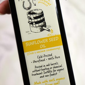 K&ouml;nnte beinhalten: Schwarze Flasche Sonnenblumen&ouml;l mit cremefarbenem Etikett. Das Etikett zeigt Illustrationen von Sonnenblumen, einem Hufeisen und einem Fass. Text: "Sonnenblumen&ouml;l", "Kaltgepresst" und "250 ml".