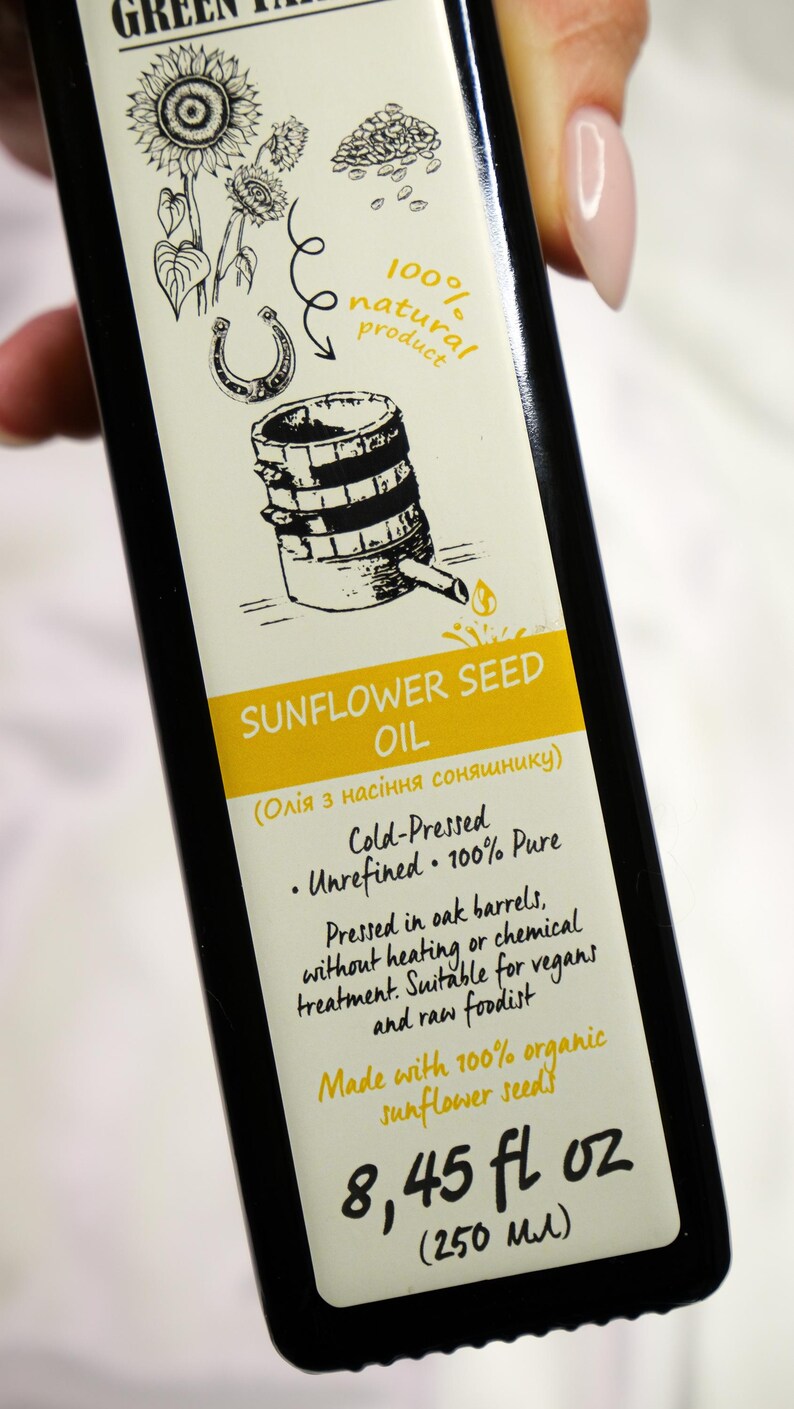 K&ouml;nnte beinhalten: Schwarze Flasche Sonnenblumen&ouml;l mit cremefarbenem Etikett. Das Etikett zeigt Illustrationen von Sonnenblumen, einem Hufeisen und einem Fass. Text: "Sonnenblumen&ouml;l", "Kaltgepresst" und "250 ml".