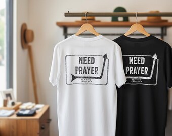 Puede incluir: Dos camisetas, una blanca y otra negra, cuelgan de perchas de madera. Las camisetas muestran el texto "NEED PRAYER" dentro de un marco rectangular con un diseño de flecha. Debajo está escrito "TAP THIS SHOULDER".