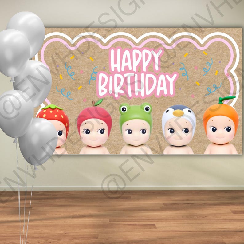 May include: A rectangular backdrop featuring the text "HAPPY BIRTHDAY" in pink, surrounded by five small figures wearing fruit and animal hats, including strawberry, apple, frog, penguin, and orange designs, with a cluster of white balloons.