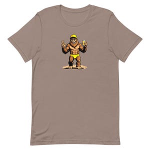 May include: A tan t-shirt featuring a cartoon illustration of a Bigfoot character wearing a yellow cap and briefs, holding a beer and giving a peace sign. The Bigfoot is brown and muscular, standing on a sandy surface.
