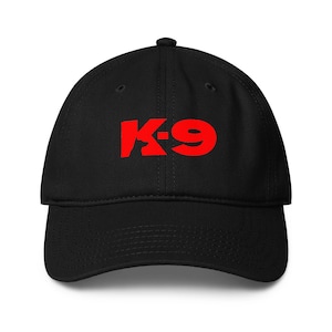 May include: Black baseball cap with a curved brim. The cap features the red text "K-9" across the front. The hat is made of a soft material and has a structured crown.