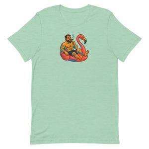 Può includere: T-shirt verde chiaro con un'illustrazione a cartoni animati di un uomo con la barba che fuma un sigaro mentre galleggia su un galleggiante a forma di fenicottero rosa. L'uomo indossa pantaloncini neri e ha tatuaggi. Il fenicottero ha dettagli arancioni e gialli.