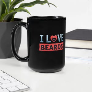Peut inclure: Mug en céramique noire avec l'inscription "I LOVE BEARDS" en blanc et rouge. Le mot "LOVE" présente un cœur avec un dessin de barbe. Le mug est posé sur une surface blanche avec une plante et un livre en arrière-plan.