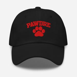 May include: Black baseball cap with the word "PAWDRE" embroidered in red above a red paw print. The cap has a curved brim and a button on top. The hat is made of a soft material.