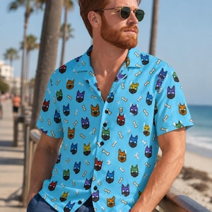 May include: A light blue short-sleeve button-down shirt featuring an all-over pattern of colorful dog faces in shades of red, green, yellow, and purple, interspersed with small white bone icons, designed with a classic open camp collar.