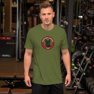 May include: Olive green t-shirt featuring a circular graphic with the words "EMOTIONAL SUPPORT" and "PUP" surrounding a black dog illustration. The shirt is short-sleeved and made of a soft material.
