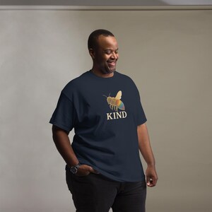 Può includere: T-shirt blu navy con una grafica di un'ape colorata e la parola "KIND" in bianco. Il design dell'ape include ali dai colori dell'arcobaleno. La maglietta è realizzata in un materiale morbido e confortevole.