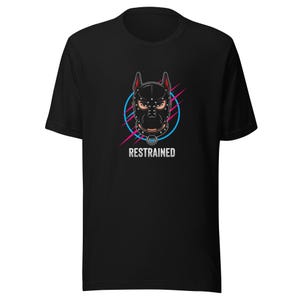 May include: A black short-sleeve t-shirt featuring a graphic of a person wearing a black studded leather-style hood with ears. The design includes a blue circle, pink diagonal lines, and the word RESTRAINED printed in white capital letters below.