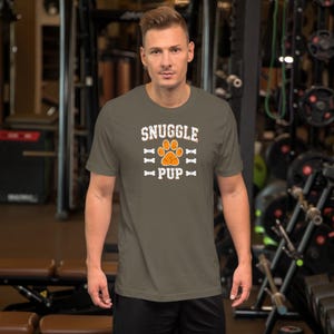 May include: Olive green t-shirt with the text "SNUGGLE PUP" in white and orange. The design includes a paw print and bone graphics. The shirt is short-sleeved and made of a soft material.