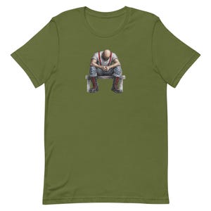 May include: An olive green t-shirt with a detailed illustration of a person sitting on a bench, head bowed. The person is wearing a white shirt, red braces, and denim jeans. The person is also wearing red boots. The image is centered on the shirt.