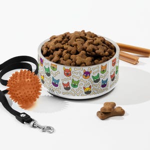 May include: A stainless steel dog bowl filled with brown kibble, featuring a colorful pattern of dog faces and bones. A black leash with an orange spiky ball and dog treats are also present, suggesting a pet-themed product.