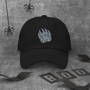 May include: Black baseball cap with a Celtic bear claw design embroidered in light gray. The cap is set against a gray background with Halloween decorations, including bats and the word "BOO".