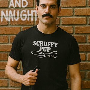 May include: Black t-shirt with the text "SCRUFFY PUP" and a leash design. The image is set against a brick wall with the words "BEARY NICE AND NAUGHTY" painted on it. The person is holding a black leash.